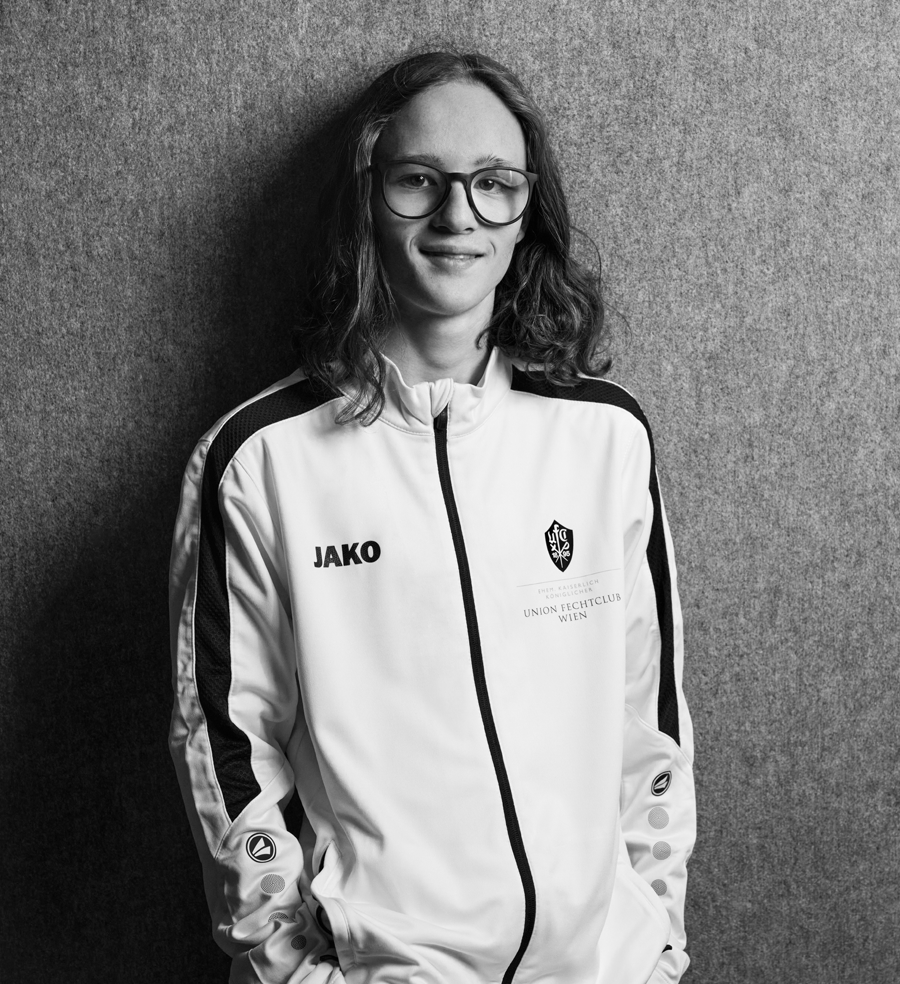 Finn, Jungbetreuer bei Partyfechten, trägt die Trainer-Weste des UFC-Wien und lächelt in die Kamera. Er betreut das Kindertraining und verfügt über einen aktuellen Erste-Hilfe-Kurs.