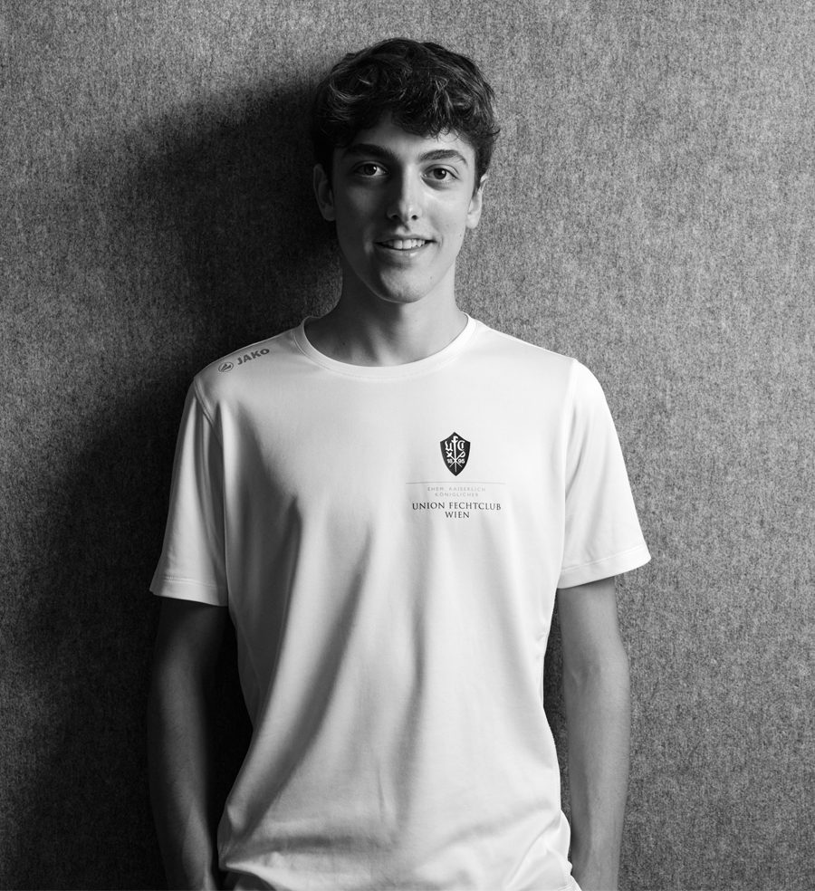 Laurenz, Jungbetreuer bei Partyfechten, trägt das Trainer-Shirt des UFC-Wien und lächelt in die Kamera. Er betreut das Kindertraining und verfügt über einen aktuellen Erste-Hilfe-Kurs.