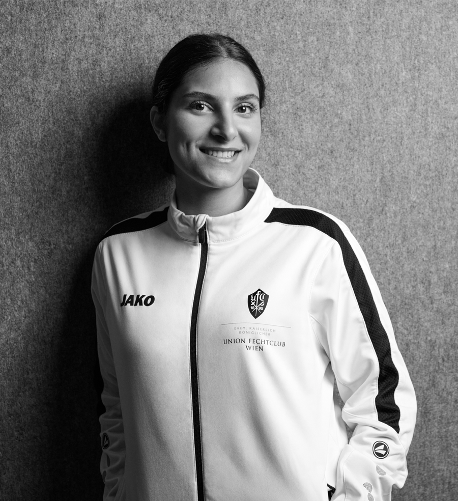 Nura, Hauptbetreuerin bei Partyfechten, trägt die Trainerweste des UFC-Wien und lächelt in die Kamera. Sie ist Fechtprofi, verfügt über langjährige Erfahrung in der Kinderbereuung und einen aktuellen Erste-Hilfe-Kurs.