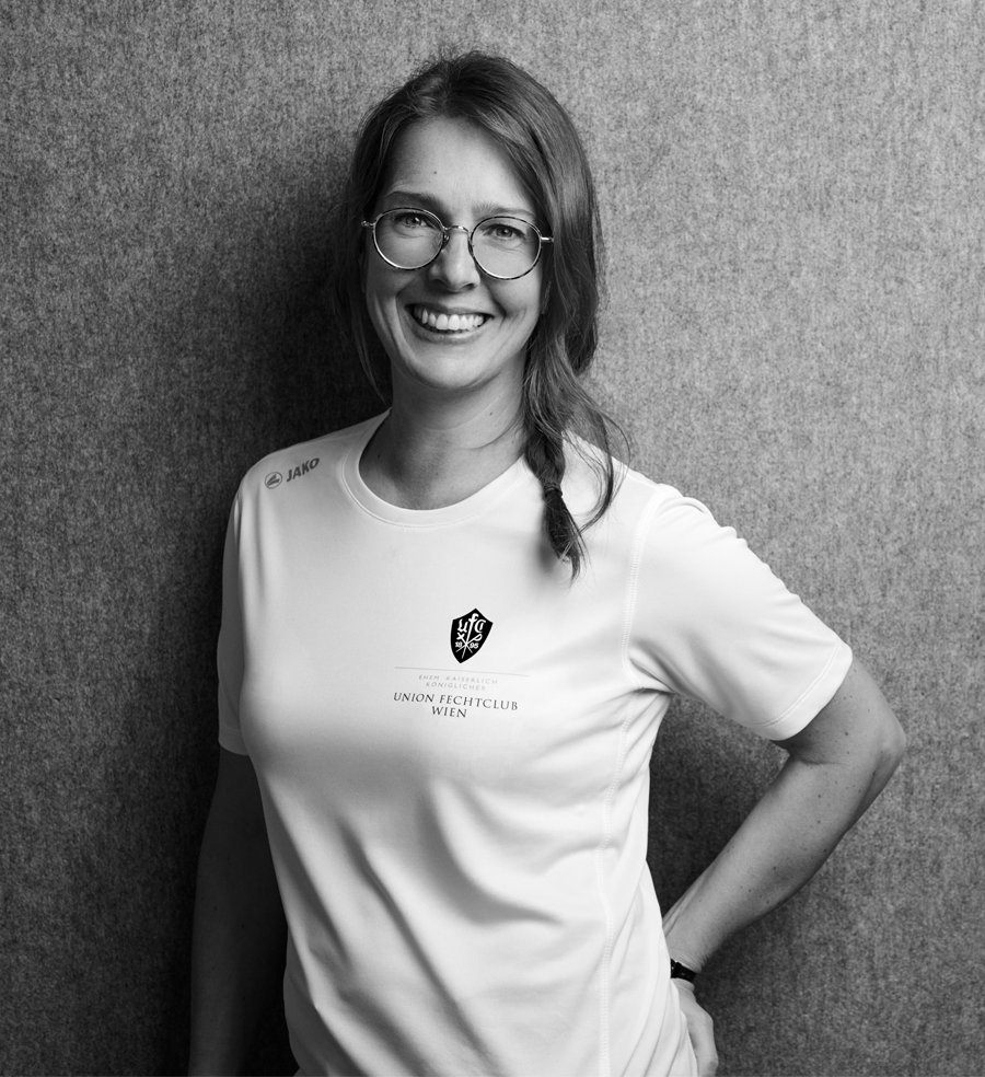 Theresa, Präsidentin des UFC-Wien und Organisatorin bei Partyfechten, trägt das Trainer-Shirt des UFC-Wien und lächelt in die Kamera. Sie ist zuständig für die reibungslosen Abläufe der Fechtpartys.