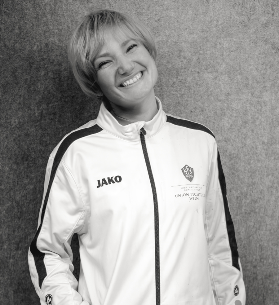 Maria, Hauptbetreuerin bei Partyfechten, trägt die Trainer-Weste des UFC-Wien und lächelt in die Kamera. Sie betreut das Kinder- und Erwachsenentraining und verfügt über einen aktuellen Erste-Hilfe-Kurs.